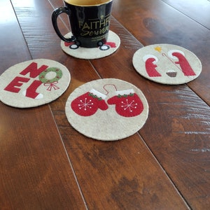 May include: Four round felt coasters with different Christmas designs. One coaster features the word "NOEL" with a wreath, another has two red mittens, one has a nativity scene, and the last one has a red truck with the words "Faithful Servant".