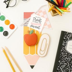 May include: Overhead shot of school-themed treats and supplies. An apple-shaped cookie with a green leaf and a paperclip-shaped cookie are next to a pencil-shaped card. The card has the text "A LITTLE TREAT TO MAKE THE FIRST DAY SWEET".