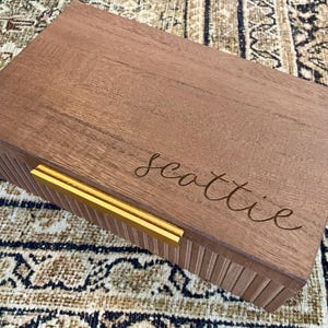 May include: A rectangular wooden box with a light brown finish. The lid is engraved with the name "Scottie" in cursive script. The box has a gold-colored handle.