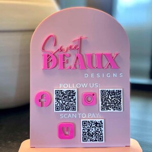 May include: A light pink arched acrylic sign featuring the text Sweet Beaux Designs in raised pink lettering. It includes three white square QR codes, a pink Facebook icon, a pink Instagram icon, and a pink payment icon with the text Follow Us and Scan To Pay.