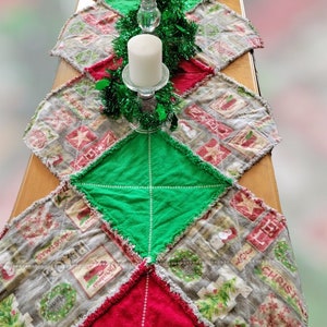May include: A red and green Christmas table runner with a patchwork design. The runner is made of fabric scraps and features a variety of Christmas-themed prints. Two white candles are placed on top of the runner, surrounded by green garland.