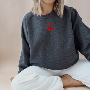 May include: A charcoal gray sweatshirt featuring a small, embroidered red cherry design on the chest. The sweatshirt has a crew neck and long sleeves. The model is wearing white pants.