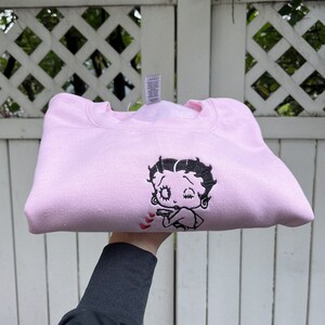 May include: A pink sweatshirt with a black embroidered image of Betty Boop blowing a kiss. The image is surrounded by small red hearts.