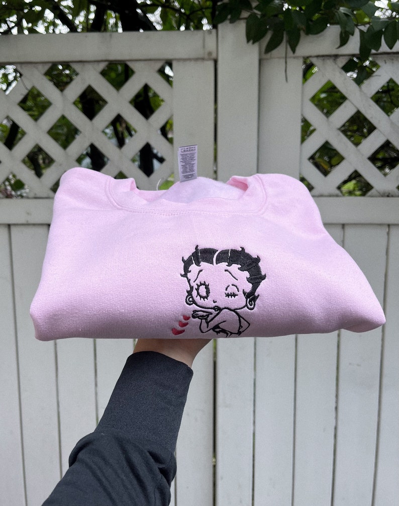 May include: A pink sweatshirt with a black embroidered image of Betty Boop blowing a kiss. The image is surrounded by small red hearts.