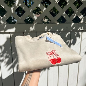 May include: A beige sweatshirt with a red cherry embroidery design. The cherries are detailed with a red bow. A blue tag is attached to the sweatshirt. The sweatshirt is folded and held up.