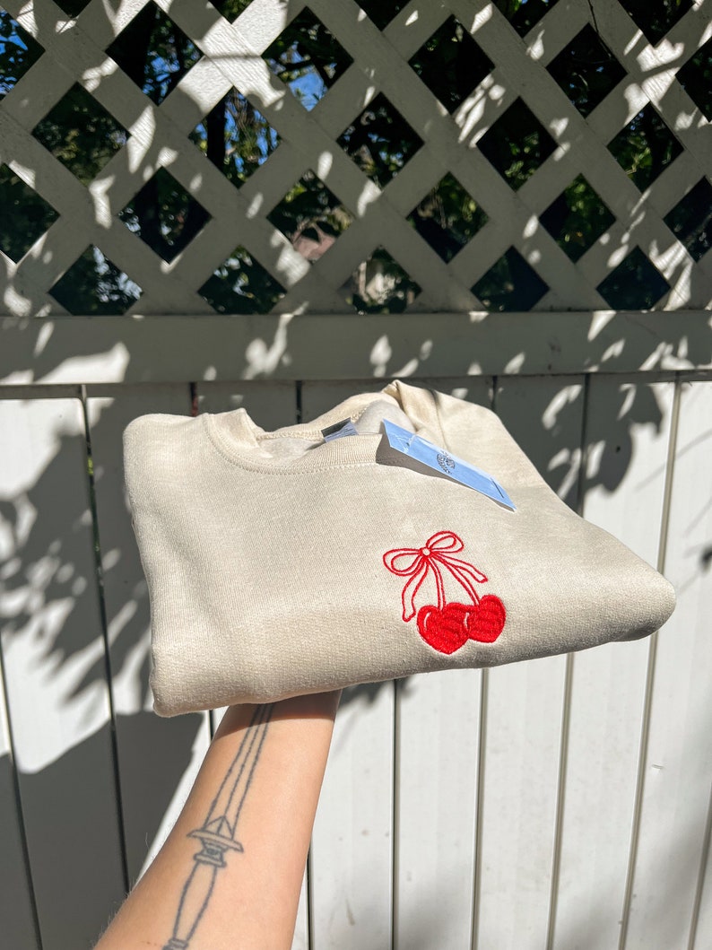 May include: A beige sweatshirt with a red cherry embroidery design. The cherries are detailed with a red bow. A blue tag is attached to the sweatshirt. The sweatshirt is folded and held up.