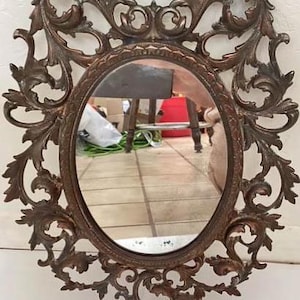 May include: Oval-shaped bronze mirror with ornate, swirling floral and leaf detailing. The mirror's frame has a dark, antique finish. The mirror reflects a room with a table and other objects.