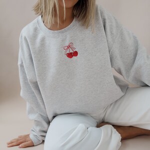 May include: A light gray sweatshirt with a crew neck and long sleeves. The sweatshirt features a small embroidered design of two red cherries with a red bow on the upper left chest. The model is wearing white pants.