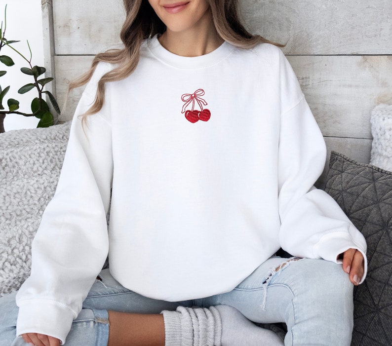 May include: White sweatshirt with a small embroidered design of two red cherries tied together with a red bow. The sweatshirt has long sleeves and a crew neck. The cherries are detailed with red stitching.