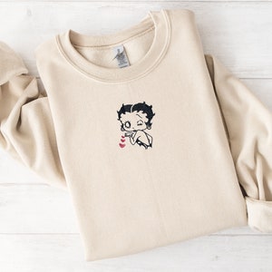 May include: A beige sweatshirt with an embroidered image of Betty Boop with hearts.
