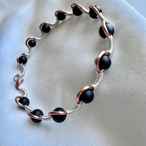 May include: A handcrafted bracelet with matte black beads, copper details, and silver-coloured wire. The beads are spaced along the wire, forming a wavy pattern. The bracelet is presented on a white surface.