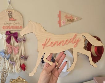 May include: Wooden horse silhouette with the name "Kennedy" written in pink on it. The horse is light brown and has a pink mane and tail.