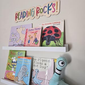 May include: A wooden sign with the words "READING ROCKS!" in colorful letters.  The sign is mounted on a white wall above a shelf with a variety of children's books, including "Bark, George!" by Jules Feiffer, "Llama Llama I Love You" by Anna Dewdney, "The Grouchy Ladybug" by Eric Carle, "Pete the Cat: Should I Share My Ice Cream?" by James Dean, and "The Pigeon Finds a Hot Dog!" by Mo Willems.  A blue plush pigeon toy is on the shelf.