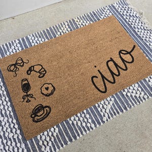 May include: A rectangular coir doormat with the word "ciao" in black script. The mat features black line drawings of a bow, croissant, wine glass, cookie, and coffee cup. The mat is on a blue and white striped rug.