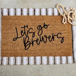 Puede incluir: Un felpudo rectangular de fibra de coco con un centro color canela que presenta el texto negro Let's go Brewers. La alfombra está colocada sobre una base de tela a rayas blancas y negras y decorada con cuentas de madera clara.