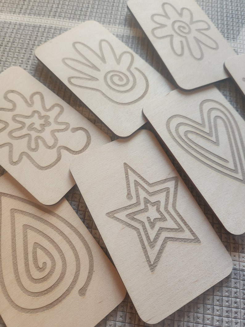 May include: Set of six wooden cards with different line art designs, including a hand, a flower, a heart, a star, a teardrop, and a swirl. The cards are all the same size and shape, and they are made of light brown wood.