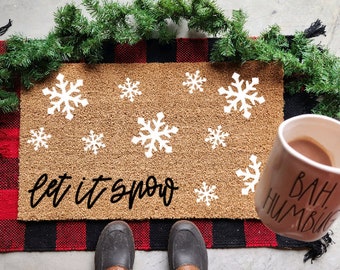 Op de afbeelding: Een rechthoekige deurmat van natuurlijke kokosvezel met witte sneeuwvlokken en de tekst "let it snow" in zwarte letters. De mat ligt op een rood-zwarte geblokte vloerkleed, met een groene slinger en een mok met "Bah Humbug".