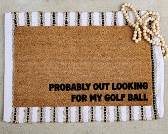 May include: A rectangular coir doormat with the text "PROBABLY OUT LOOKING FOR MY GOLF BALL". The mat is brown with a white border and black and beige stripes. A strand of wooden beads rests on the mat.