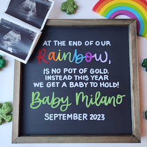 May include: A chalkboard sign with a wooden frame announcing a pregnancy with the text "At the end of our rainbow, is no pot of gold. Instead this year we get a baby to hold! Baby Milano September 2023".