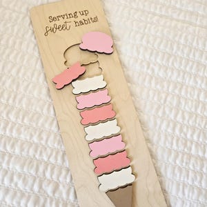 以下が含まれることがあります： ピンク、白、ベージュの層を持つ木製のアイスクリームコーンパズル。「Serving up sweet habits!」の文字が入った明るい木製のベースにパズルが配置されています。パズルのピースは取り外し可能です。