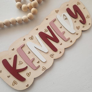 May include: A wooden sign with the name "KENNEDY" spelled out in red, pink, and white letters. The letters are surrounded by small gold hearts.