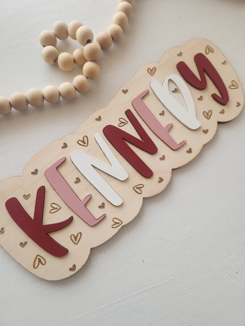 May include: A wooden sign with the name "KENNEDY" spelled out in red, pink, and white letters. The letters are surrounded by small gold hearts.