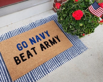 Può includere: Un tappetino marrone con la scritta "GO NAVY BEAT ARMY" in lettere blu e nere. Il tappetino è su un tappeto a strisce blu e bianche. Una pianta in vaso con fiori rossi è a destra del tappetino.
