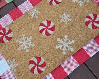 May include: A rectangular coir doormat with a festive design featuring red and white peppermint candies and snowflakes. The mat is set on a red and white checkered fabric, perfect for holiday decor.