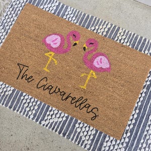May include: A brown coir doormat with a white and blue striped border. The doormat features two pink flamingos and the text "The Ciavarellas".