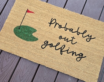 May include: A brown coir doormat with the text "Probably out golfing" in black cursive font. A green golf green with a red flag is in the top left corner.
