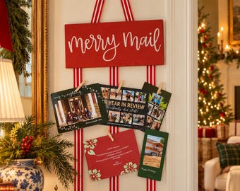 May include: A red wooden sign with the white text merry Mail hanging from a gold hook by red and white striped ribbons. Three vertical red and white ribbons are attached to the sign, holding four holiday cards with wooden clothespins.
