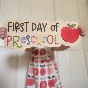 May include: A child holds a wooden sign that says "FIRST DAY OF PRESCHOOL" with a red apple graphic. The sign is decorated with colorful letters.