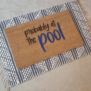 May include: A rectangular coir doormat with the phrase "probably at the pool" in black and blue lettering, placed on a striped rug with fringe. The rug has a white and blue pattern.