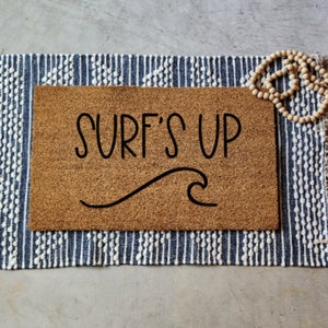 以下が含まれることがあります： 「SURF'S UP」の黒い文字と波のグラフィックが描かれた長方形のコイアのドアマット。マットは、フリンジ付きの青と白のストライプのラグの上に置かれ、木製のビーズでアクセントがつけられています。