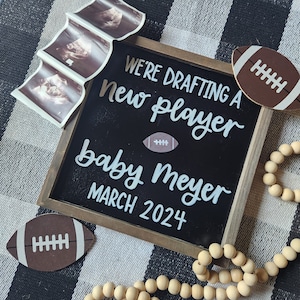 May include: A black chalkboard sign with white lettering announcing "We're drafting a new player, baby Meyer, March 2024". The sign is surrounded by brown footballs, a wooden bead garland, and three ultrasound images.