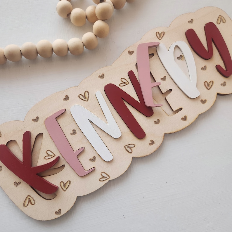 May include: A wooden sign with the name "KENNEDY" spelled out in pink, white, and red letters. The letters are cut out and layered on top of each other. The sign is decorated with small hearts.