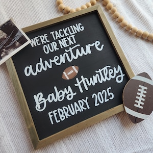 May include: A black chalkboard sign with a white wooden frame announces "We're tackling our next adventure Baby Huntley February 2025". A football graphic is next to the text.