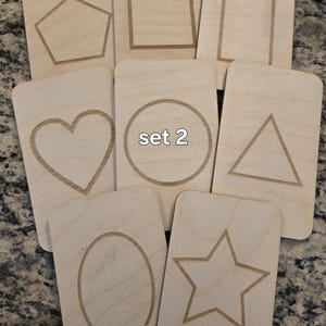 May include: Set of eight wooden cards with different shapes cut out. The shapes include a heart, circle, triangle, star, oval, square, rectangle, and pentagon. The cards are labeled "set 2".