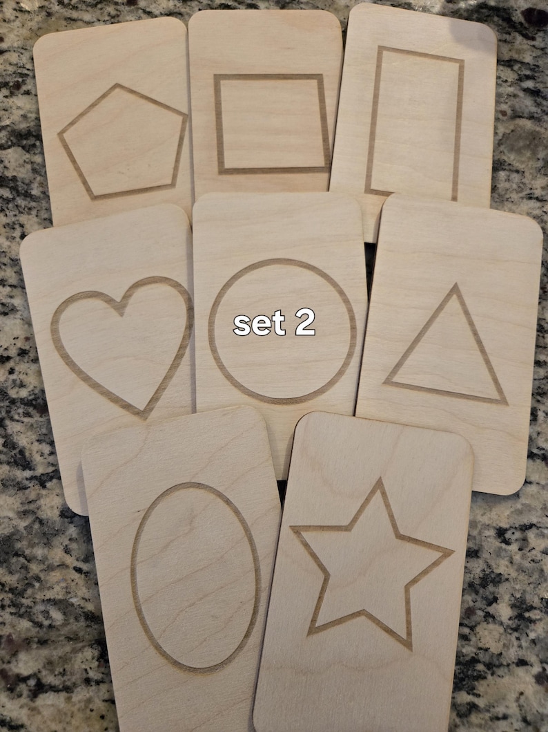 May include: Set of eight wooden cards with different shapes cut out. The shapes include a heart, circle, triangle, star, oval, square, rectangle, and pentagon. The cards are labeled "set 2".