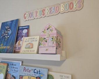 Puede incluir: Un letrero de madera con el texto "COOL KIDS READ BOOKS" en colores pastel cuelga sobre una estantería llena de libros infantiles. Una caja de joyería rosa floral se encuentra en la estantería, junto con varios títulos de libros.