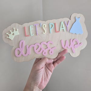 May include: A wooden sign with the words "Let's Play Dress Up" in colorful letters. A crown and a blue dress are also on the sign.