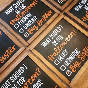 May include: Wooden framed black chalkboard signs with white and orange text. The signs ask "WHAT SHOULD I BE FOR Halloween?" with costume options like "Fireman", "Dinosaur", and "Big Brother".