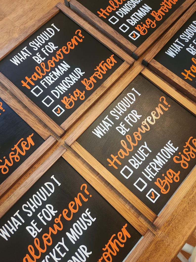 May include: Wooden framed black chalkboard signs with white and orange text. The signs ask "WHAT SHOULD I BE FOR Halloween?" with costume options like "Fireman", "Dinosaur", and "Big Brother".