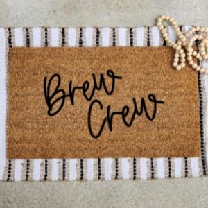 Puede incluir: Un felpudo rectangular de fibra de coco marrón sobre una alfombra de tela a rayas blancas y negras. El felpudo presenta el texto negro Brew Crew en letra cursiva. Un collar de cuentas de madera clara descansa en la esquina superior.