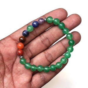 May include: A gemstone bracelet with a mix of coloured beads. The bracelet features red, orange, brown, blue, purple, and green beads. The green beads are a consistent size, while the other beads vary. The bracelet is held in a hand.