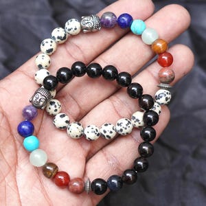 May include: Two beaded bracelets with a variety of colourful stones and black beads. One bracelet features a silver Buddha head charm. The bracelets are held in a hand against a dark background. The beads are various sizes and colours.