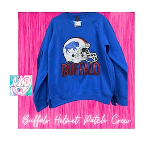 May include: A royal blue sweatshirt with a football helmet graphic. The helmet is white with a blue buffalo logo and the word "BUFFALO" in red. The text "Buffalo Helmet Patch Crew" is below the sweatshirt.