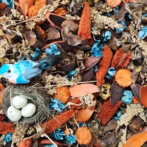 Könnte beinhalten: Eine blaue Vogelfigur, die auf einem braunen geflochtenen Nest mit drei weißen Eiern sitzt. Das Nest ist umgeben von einer Vielzahl getrockneter Blumen, Blätter und Hülsen in Braun-, Orange- und Blautönen.