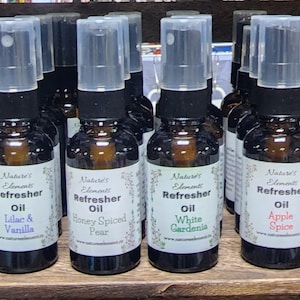 Puede incluir: Una fila de pequeñas botellas de vidrio con pulverizadores, cada una llena de un aceite perfumado diferente. Las botellas están etiquetadas con el texto "Nature's Element Refresher Oil" y el nombre del aroma, como "Lilac & Vanilla", "Honey Spiced Pear", y "White Gardenia".