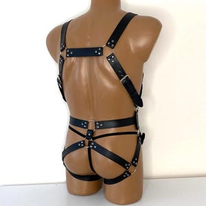 Op de afbeelding: Zwart leren bodyharnas met verstelbare bandjes, metalen ringen en gespen. Het harnas heeft een borstband, schouderbanden en taillebanden, verbonden door metalen ringen. Het ontwerp bevat een string-stijl onderkant.